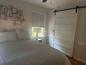 Ein Schlafzimmer mit einem Bett, einem Fenster mit Jalousien, einem Nachttisch und einer Schiebetür.