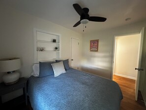 A bedroom with a bed, a ceiling fan, a nightstand, a lamp, a shelf with decorative items, and a door. - Quiet Denver Home Near DU, Cherry Creek, Wash Park & Wellshire Golf (Denver)