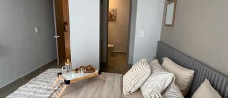 Un dormitorio moderno con cama, mesitas de noche, una lámpara y un baño visible a través de una puerta abierta.