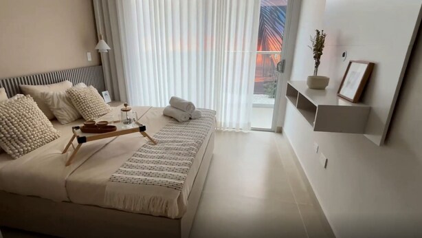 A modern bedroom with a bed, pillows, a bedside table with a tray, a potted plant, and a view of a bridge through the window. - Praia Suites by Avia Suites (Santa Marta)