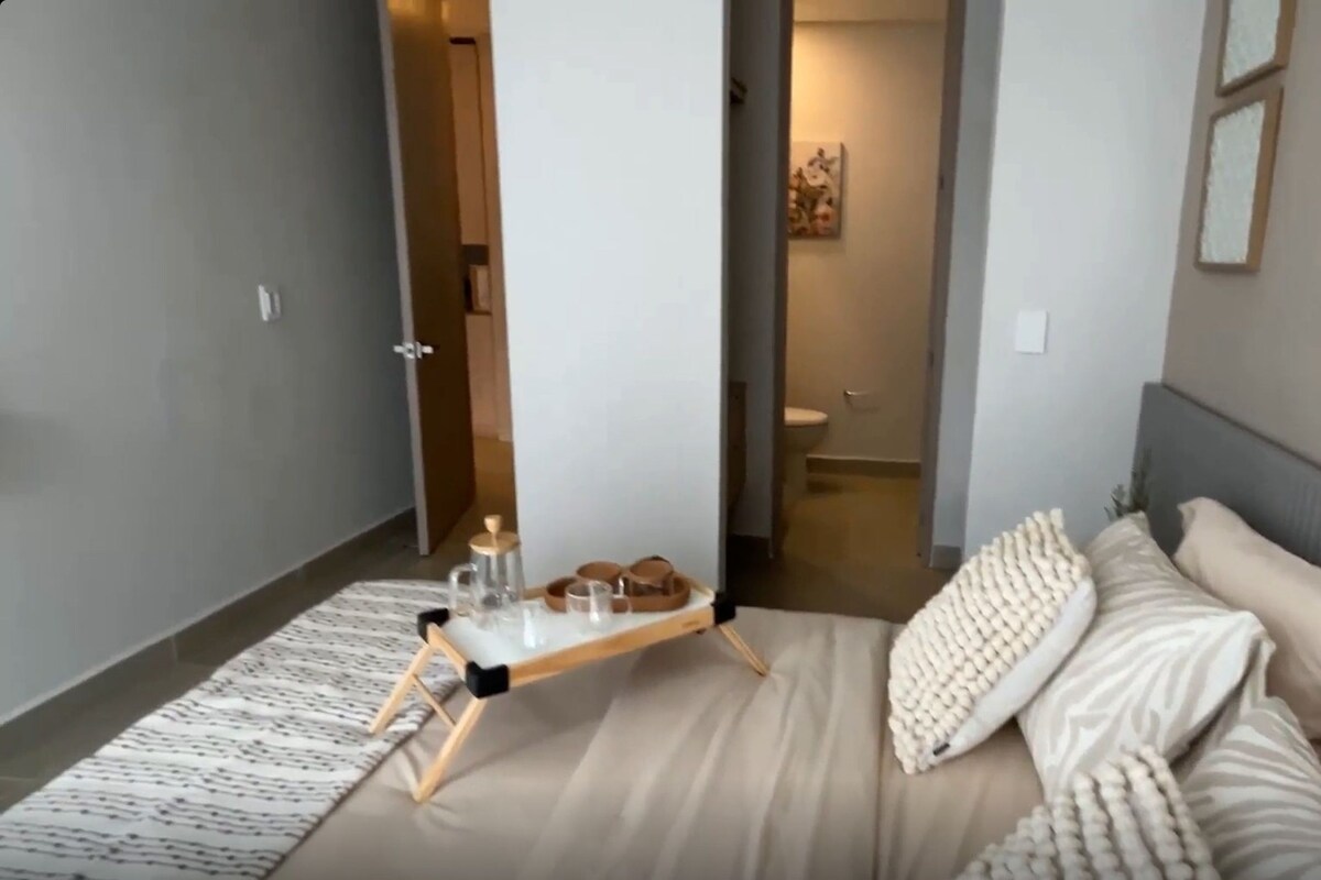 Un dormitorio moderno con cama, mesitas de noche, una lámpara y un baño visible a través de una puerta abierta.