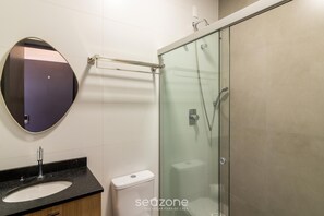 A bathroom with a glass-enclosed shower, a round mirror, and a sink with a black countertop. - Studio in Condo w/ Parking in Blumenau Ola0004 (Blumenau)