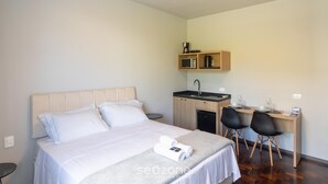 Quarto de hotel com uma cama, uma secretária com duas cadeiras, uma kitchenette pequena e uma mesa de cabeceira.