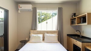A hotel room with a bed, a small bedside table, a built-in microwave, a coffee maker, and a window with curtains. - Studio 750m From Vila Germanica Ola0005 (Blumenau)