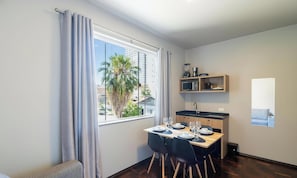 A compact kitchen with a microwave, sink, and wooden shelves. A dining table with chairs is set up near a window with curtains. - Studio w/ Garage, 750m fr/ Vila Germanica Ola0006 (Blumenau)