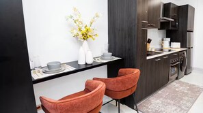 A modern kitchen with dark cabinetry, a black countertop, and two orange chairs. - Executive Studio at LoxwoodHouse, East Legon (Accra)