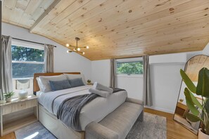 Un dormitorio con techo de madera, cama con ropa de cama gris, mesita de noche con lámpara y ventana con cortinas.