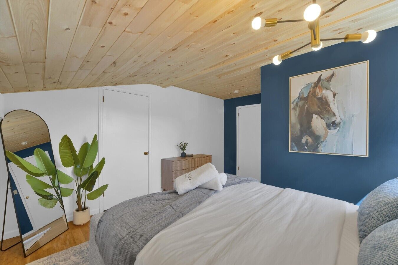 A bedroom with a bed, bedside tables, a mirror, a plant, and a painting on the wall.