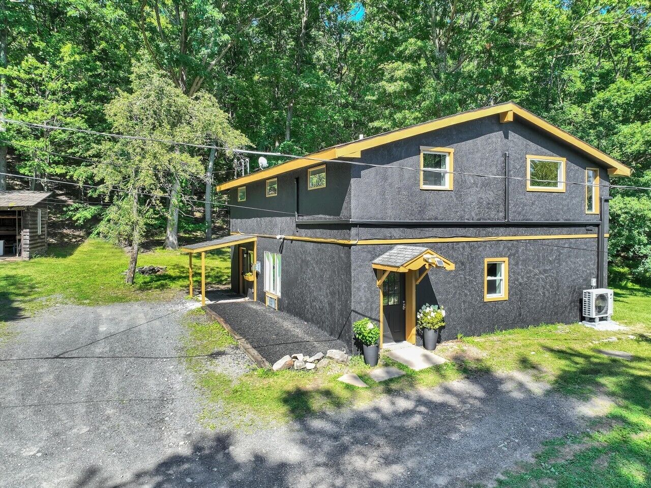 A two-story house with a dark exterior and yellow trim, a front porch, and a gravel driveway.