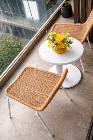 Ein Korbstuhl und ein kleiner runder Tisch mit Blumenarrangement auf einem Balkon mit Glasgeländer.