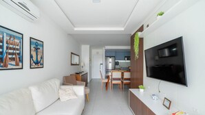A modern living room with a white sofa, a flat-screen TV mounted on the wall, and a dining area with wooden chairs and a table. - Apart w/ BBQ in Condo w/ Pool Rmm0206 (Florianópolis)