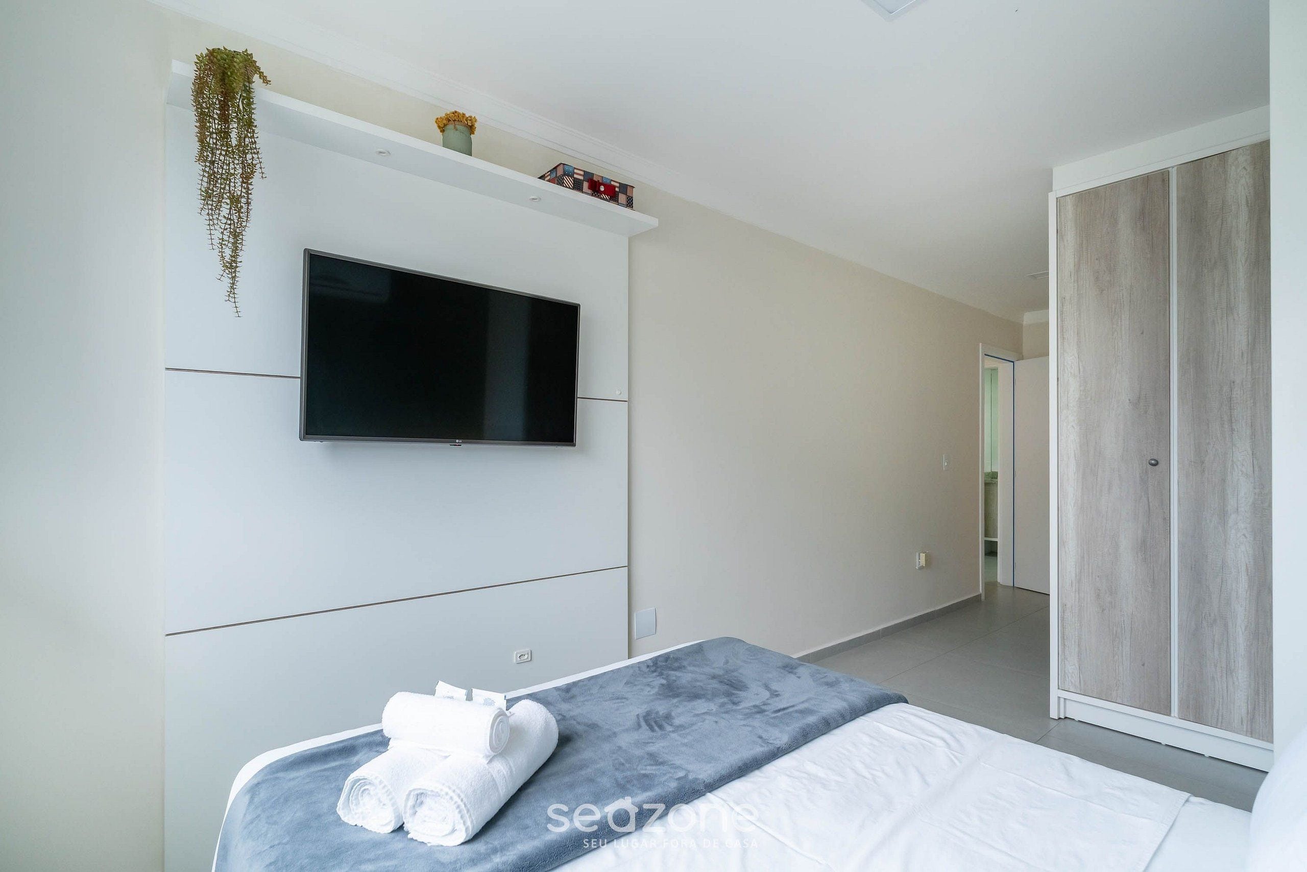 A modern bedroom with a bed, a flat-screen TV mounted on the wall, a wardrobe, and a hanging plant.