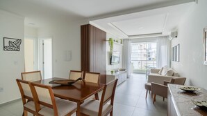 A modern living room with a dining area, a TV mounted on the wall, and a view of the city through large windows. - Apart w/ BBQ in Condo w/ Pool Rmm0206 (Florianópolis)