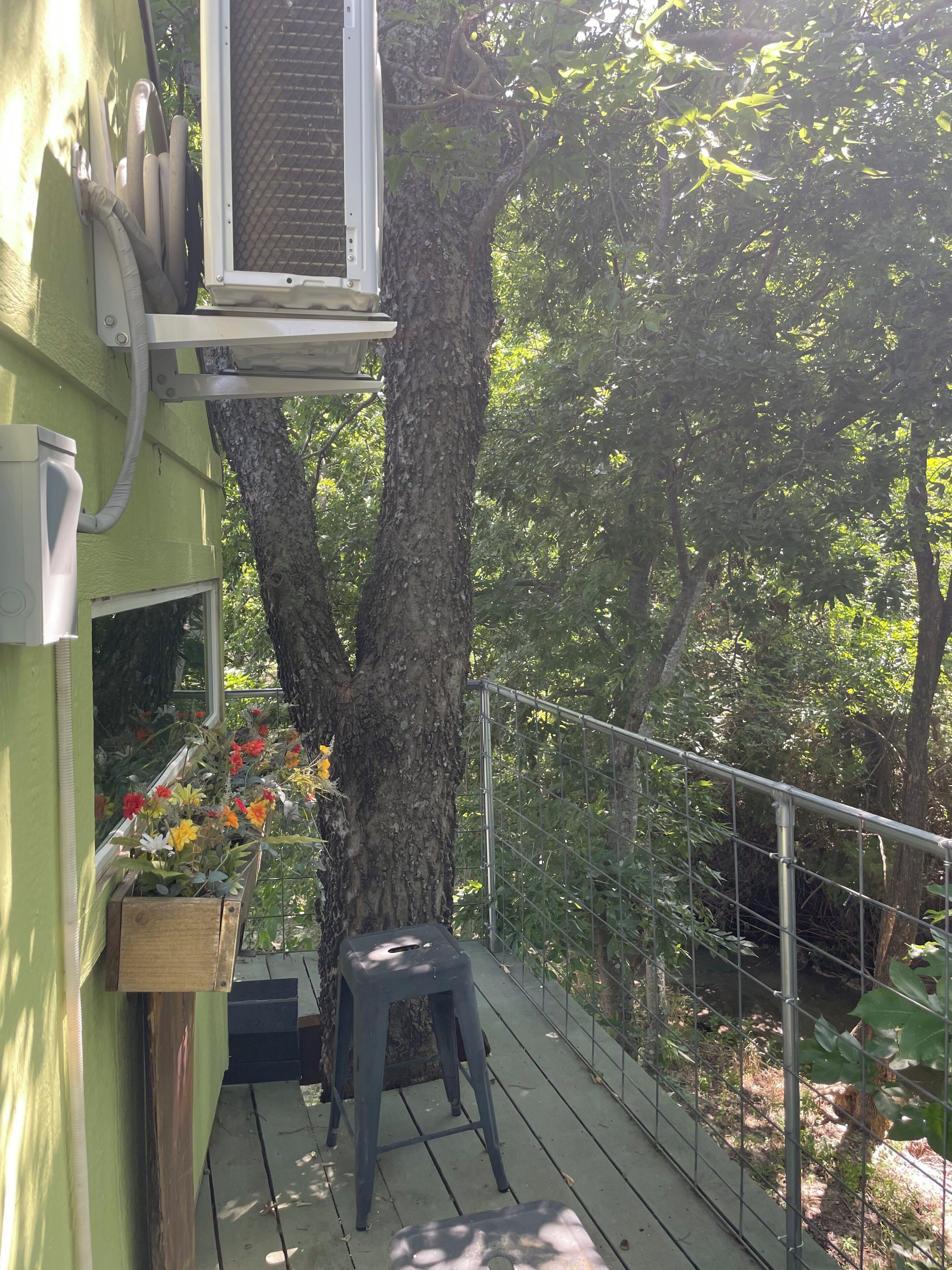 Un balcón con una jardinera de madera con flores, baranda de metal y un árbol.