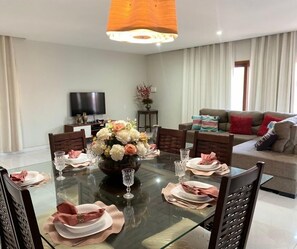 Comedor con mesa de vidrio dispuesta para seis personas, sillas, televisor y sofá con cojines.
