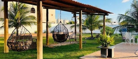 A covered outdoor seating area with hanging chairs, palm trees, and a view of the ocean.