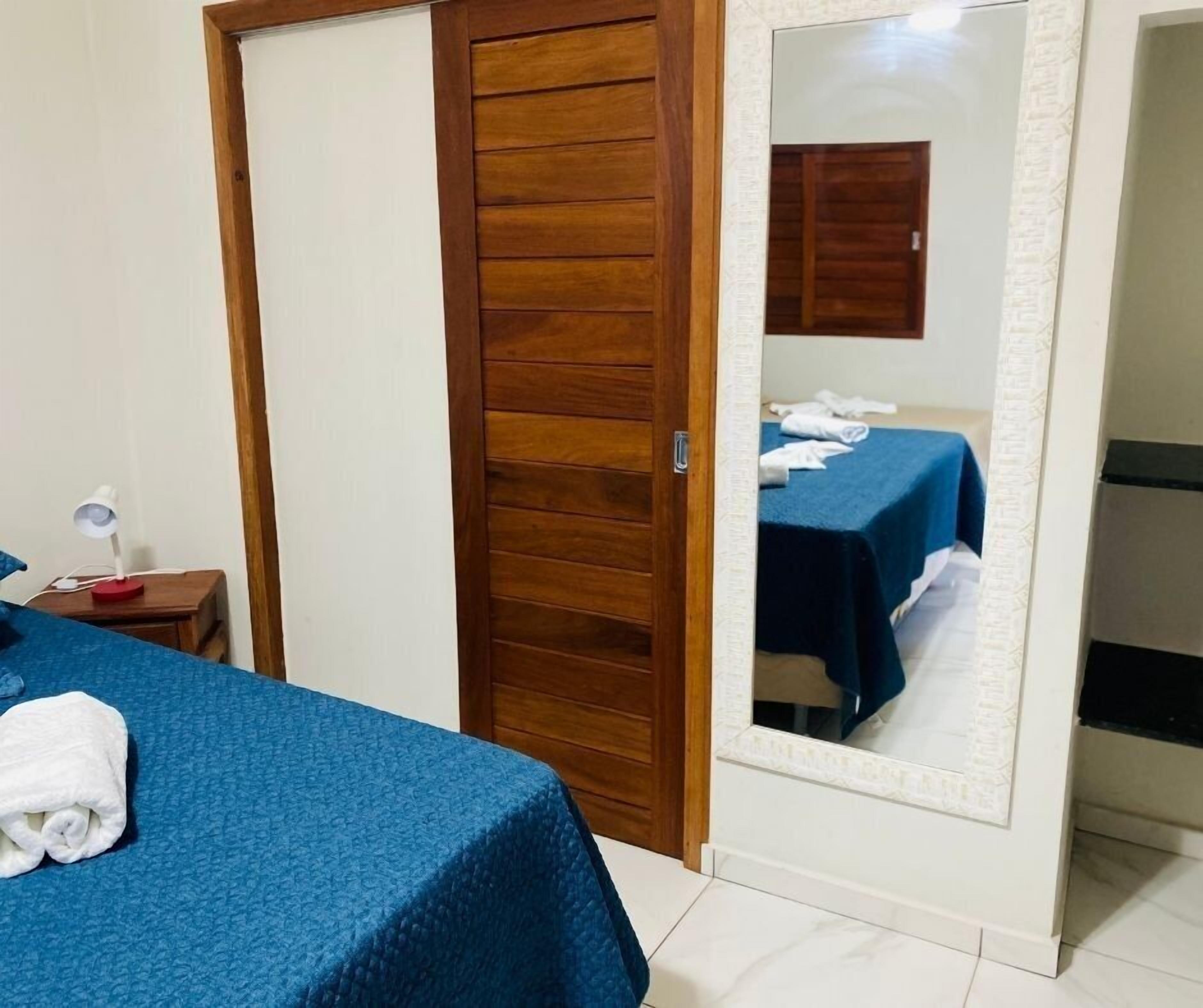 Quarto de hotel com porta de madeira, cama com colcha azul, mesa de cabeceira com abajur e um espelho refletindo o quarto.