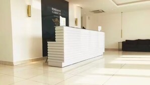 A reception desk in a hotel lobby with a tiled floor and a black wall with a logo. - Urban Studio+BigComfy Bed+EmbsyGdns, Cantonments (Accra)