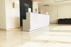 A reception desk in a hotel lobby with a tiled floor and a black wall with a logo. - Urban Studio+BigComfy Bed+EmbsyGdns, Cantonments (Accra)
