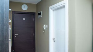 A hallway with a dark wooden door, a white door, a clock, and a phone on the wall. - Urban Studio+BigComfy Bed+EmbsyGdns, Cantonments (Accra)