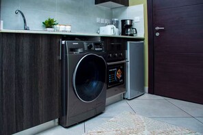 A compact kitchen with a washing machine, a coffee maker, and a small refrigerator. - Urban Studio+BigComfy Bed+EmbsyGdns, Cantonments (Accra)