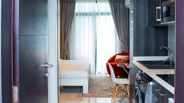A modern hotel room with a bed, a seating area with a chair, a kitchen with appliances, and a view of the outdoors. - Urban Studio+BigComfy Bed+EmbsyGdns, Cantonments (Accra)