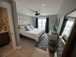 Um quarto com cama, ventilador de teto, janela com cortinas e vista para a água.