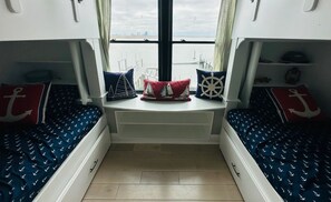 Una stanza con due letti, ognuno con una coperta blu decorata con ancora. Cuscini con motivi a ancora e barca a vela. Si vede una finestra con tende, e una piccola mensola con una lanterna a destra.