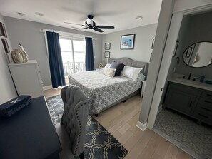 A bedroom with a bed, a desk, a chair, a ceiling fan, a window with curtains, and a bathroom with a sink and mirror. - Spacious 5-Bedroom house on the bay with large dock and open water, sunset views (Brigantine)