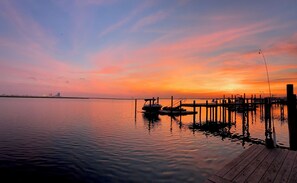 Una tranquilla riva all'ora del tramonto con un molo che si protende nell'acqua, una barca ormeggiata al molo e sullo sfondo la silhouette di una città.