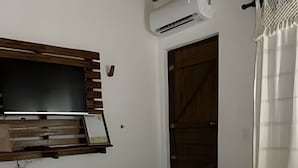 Una cama bien hecha con edredón y almohadas blancos, un televisor montado en un estante de madera y una puerta que da a una habitación oscura.