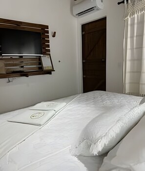 A neatly made bed with a white duvet and pillows, a mounted television on a wooden shelf, and a door leading to a dark room.