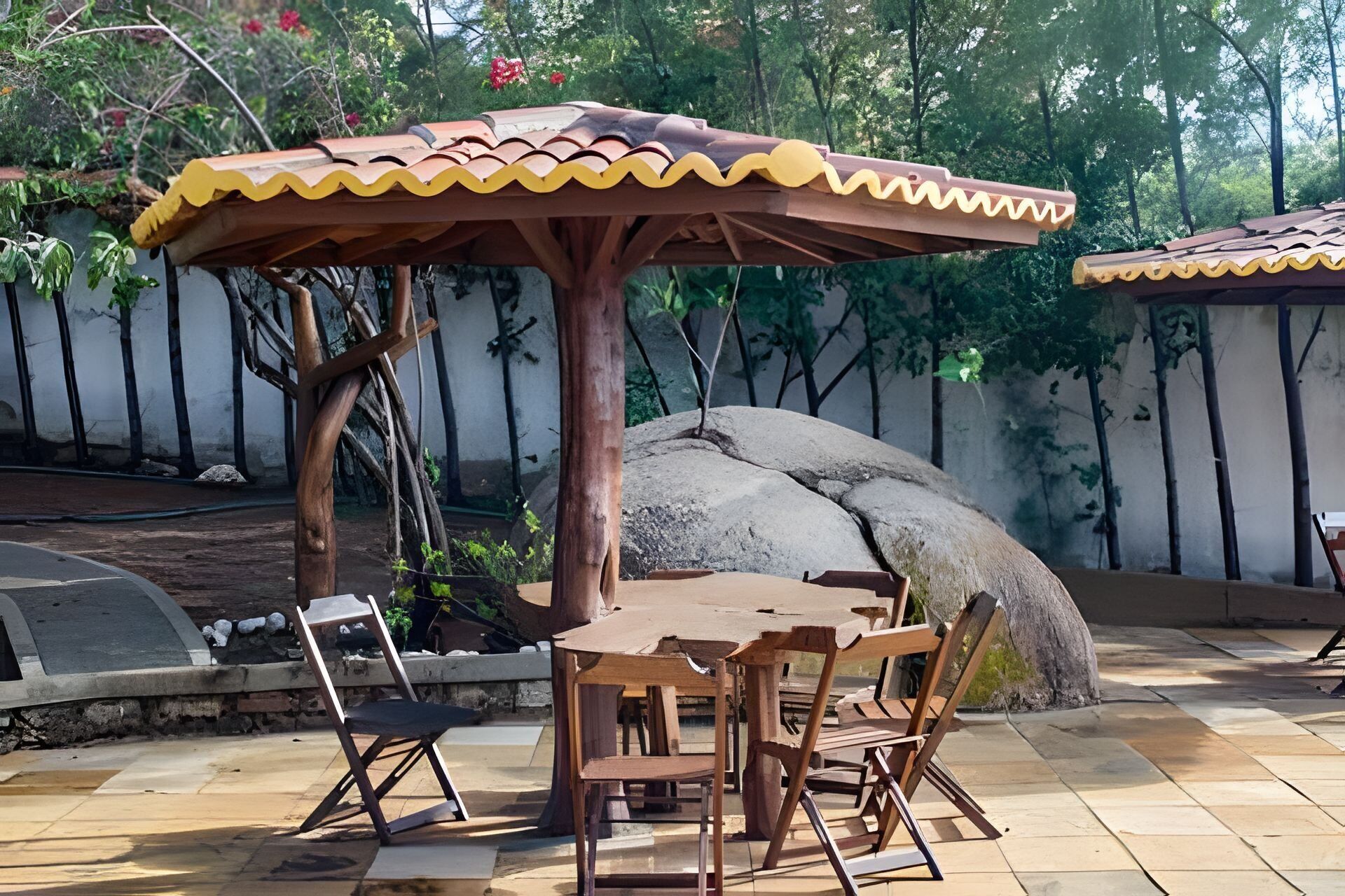 Un patio al aire libre con una mesa y sillas de madera, un paraguas decorativo y una gran roca.