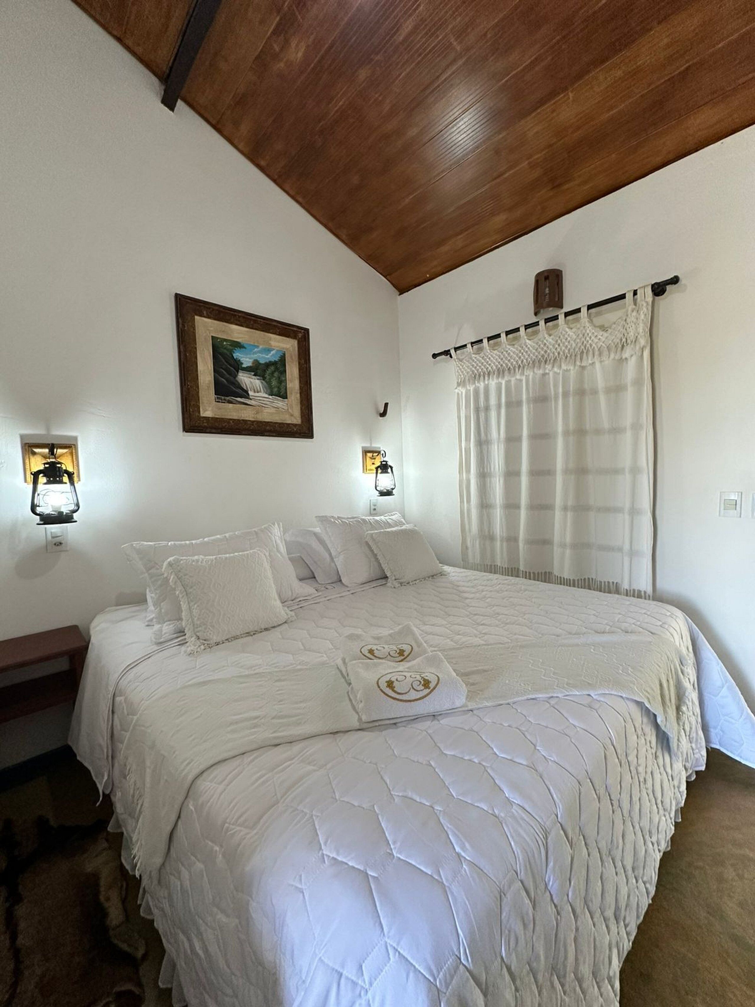 Quarto com uma cama grande, duas luminárias de cabeceira, um quadro na parede e uma janela com cortinas.