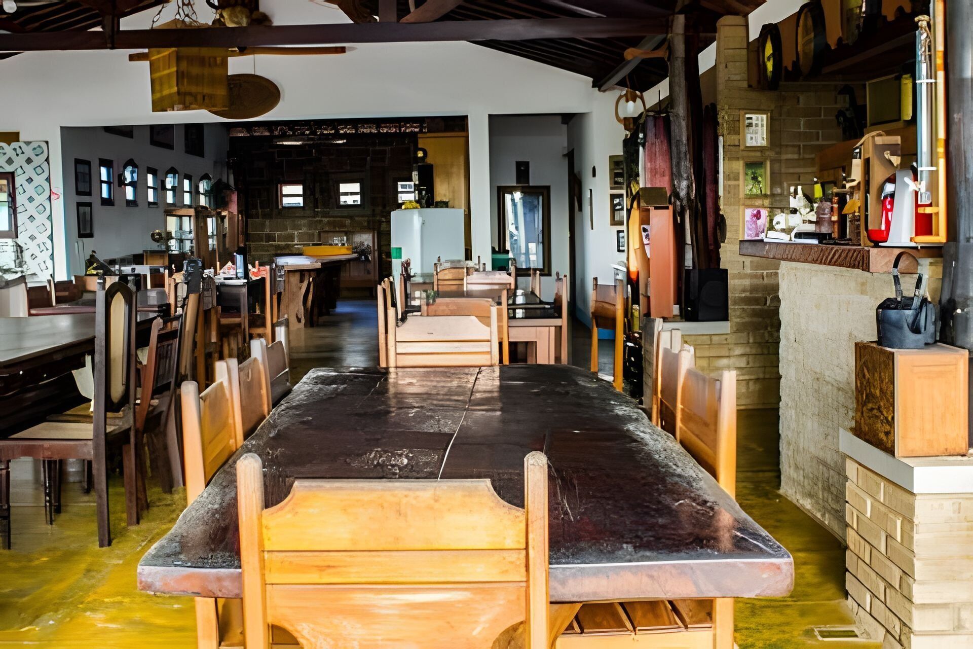 Uma sala de jantar ampla com uma mesa comprida e várias cadeiras, uma cozinha com diversos eletrodomésticos e uma parede de tijolos com prateleira.