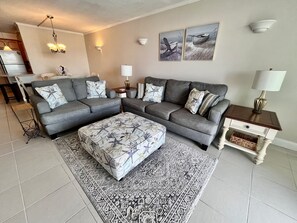 Ein Wohnzimmer mit grauem Ecksofa, einem gemusterten Hocker und einem Teppich mit Muster. An den Wänden hängen Lampen und Kunstwerke, die Boote zeigen.