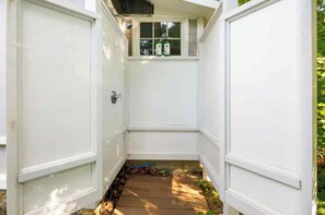 Ein schmaler Außenweg führt zu einer weißen Tür mit einem Fenster darüber, umgeben von viel Grün.