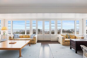 Ein geräumiges Zimmer mit großen Fenstern, einem Holztisch, zwei Sofas und einem dunklen Holzschrank.