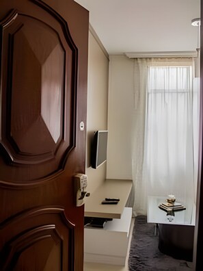 A hotel room with a wooden door, a flat-screen TV on a white stand, a small desk with a keyboard, a chair, and a window with sheer curtains. - 4let BH Del Rey (Belo Horizonte)