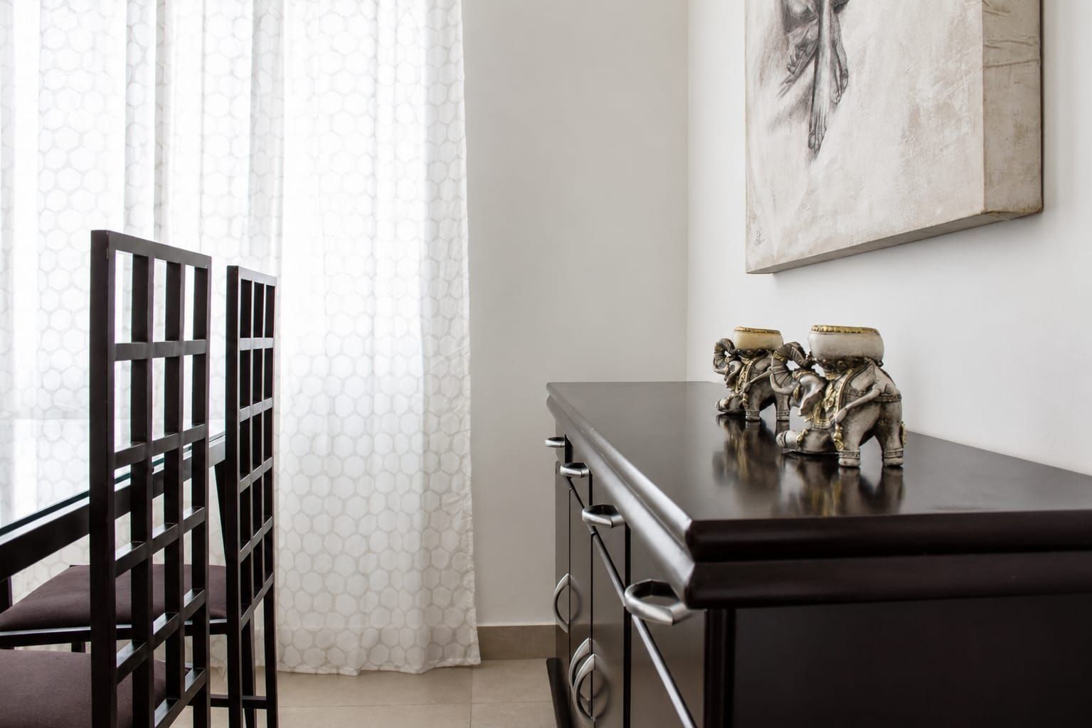 Quarto com área de jantar, apresentando uma cadeira de madeira e uma mesa, cômoda preta com objetos decorativos e um quadro na parede.