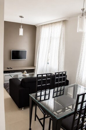 A modern living room with a glass dining table, black chairs, a black sofa, a flat-screen TV, and a coffee table with books and a vase.