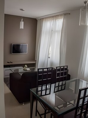 A modern dining area with a glass table and chairs, a sofa, a flat-screen TV mounted on the wall, and a window with sheer curtains. - 4let BH Del Rey (Belo Horizonte)