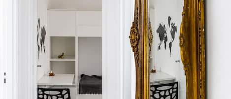 Un pasillo con un espejo grande y decorativo, una puerta blanca y una habitación con un armario blanco y una silla negra.