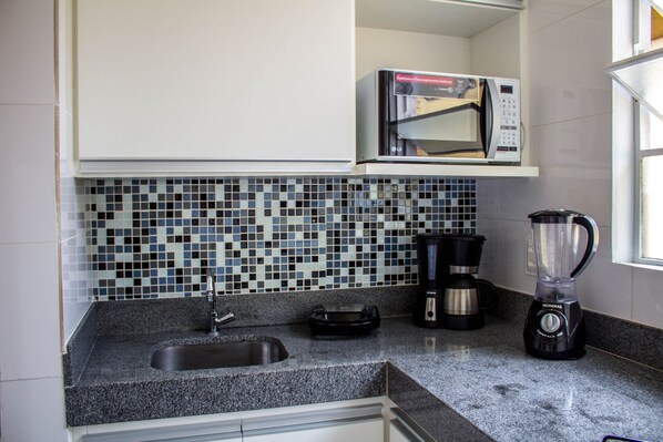 A kitchen with a granite countertop, a sink, a coffee maker, a blender, and a microwave above the tiled backsplash.