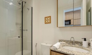 Ein modernes Badezimmer mit einer gläsernen Duschkabine, einer Toilette, einem Waschbecken mit Granitplatte und einem Spiegel, der auf einen Küchenbereich blickt.