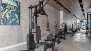 A gym with exercise equipment, a bench, and a large abstract painting on the wall. - Studio in Condo With Pool in Republica Thk1902 (São Paulo)