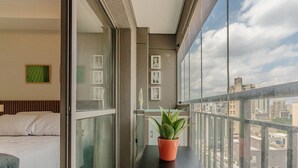 A balcony with a potted plant, a bed with white linens, and a cityscape view. - Studio in Condo With Pool in Republica Thk1902 (São Paulo)