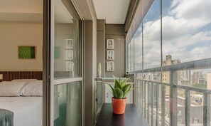 Ein Balkon mit einer Topfpflanze, ein Bett mit weißer Bettwäsche und Blick auf die Stadt.