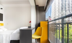 Quarto de hotel moderno com cama, mesa pequena, cadeira amarela e varanda com vista para a cidade.