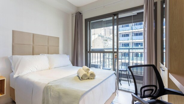 A hotel room with a bed, a chair, and a large window with a view of buildings. - Complete Studio 350m From the Metro Thk0502 (São Paulo)